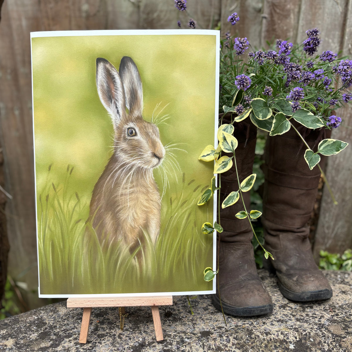 Hare In The Grass Fine Art Print On Paper on an easel outdoors beside rustic boots and lavender, perfect for farmland-themed interiors.
