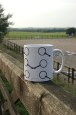 Snaffle Mug - Country Kitchenware featuring hand-drawn snaffle bit design, placed on a wooden fence with a countryside equestrian arena in the background.
