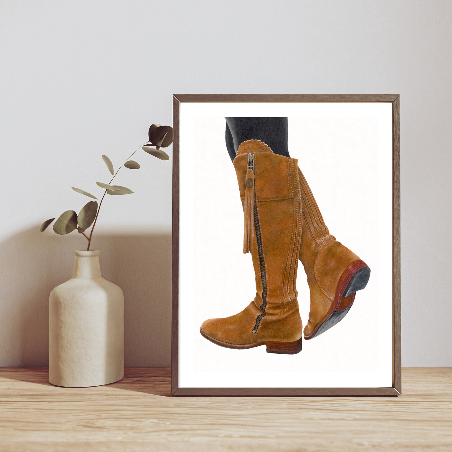 Framed picture of brown leather boots on a wooden surface with a vase and plant in the background.