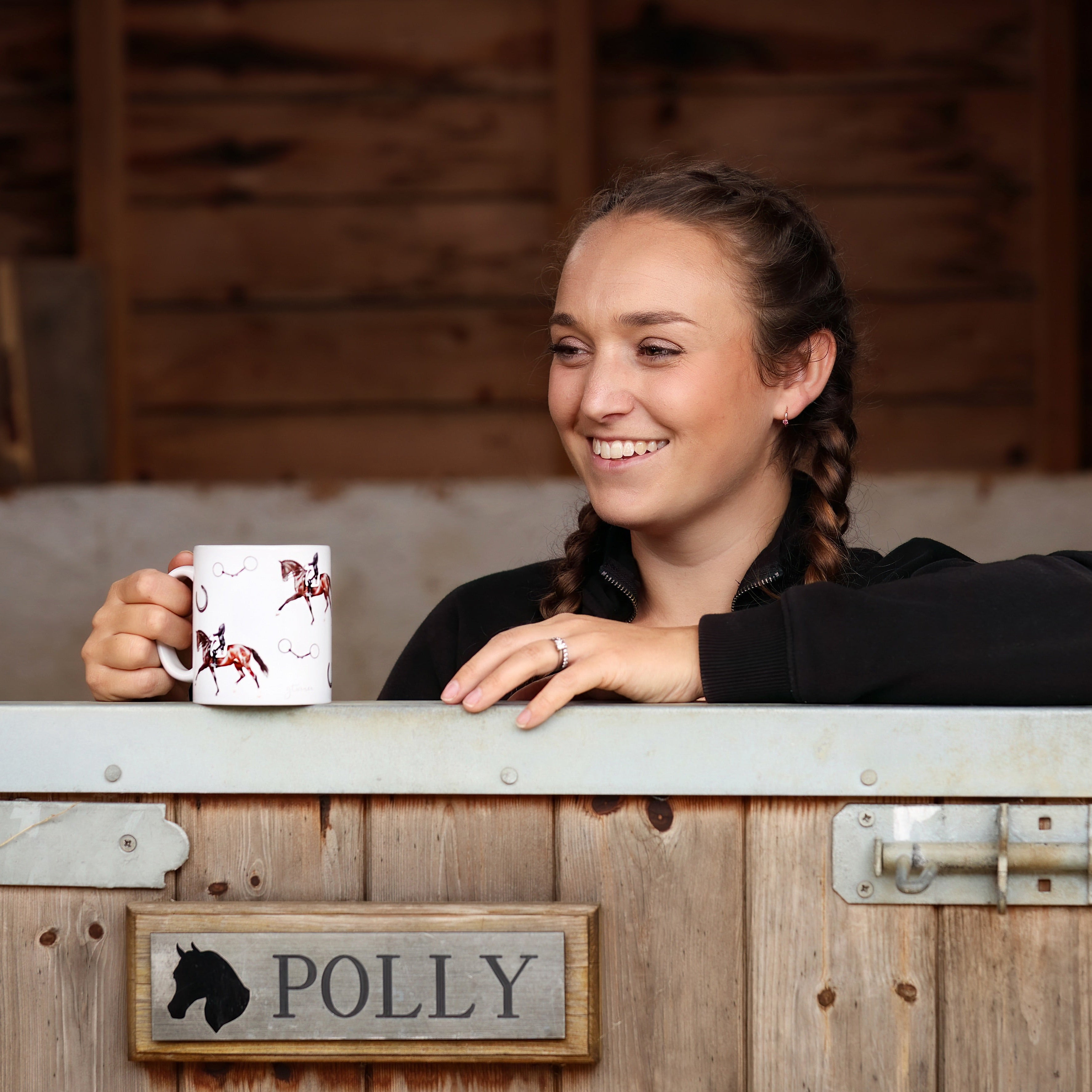 Woman holding a mug with a horse design, sitting at a wooden desk with 'Polly' sign.