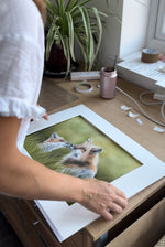 Artist preparing framed Fox & Cub original artwork on a wooden desk, highlighting fine detail and craftsmanship in country-style home decor.