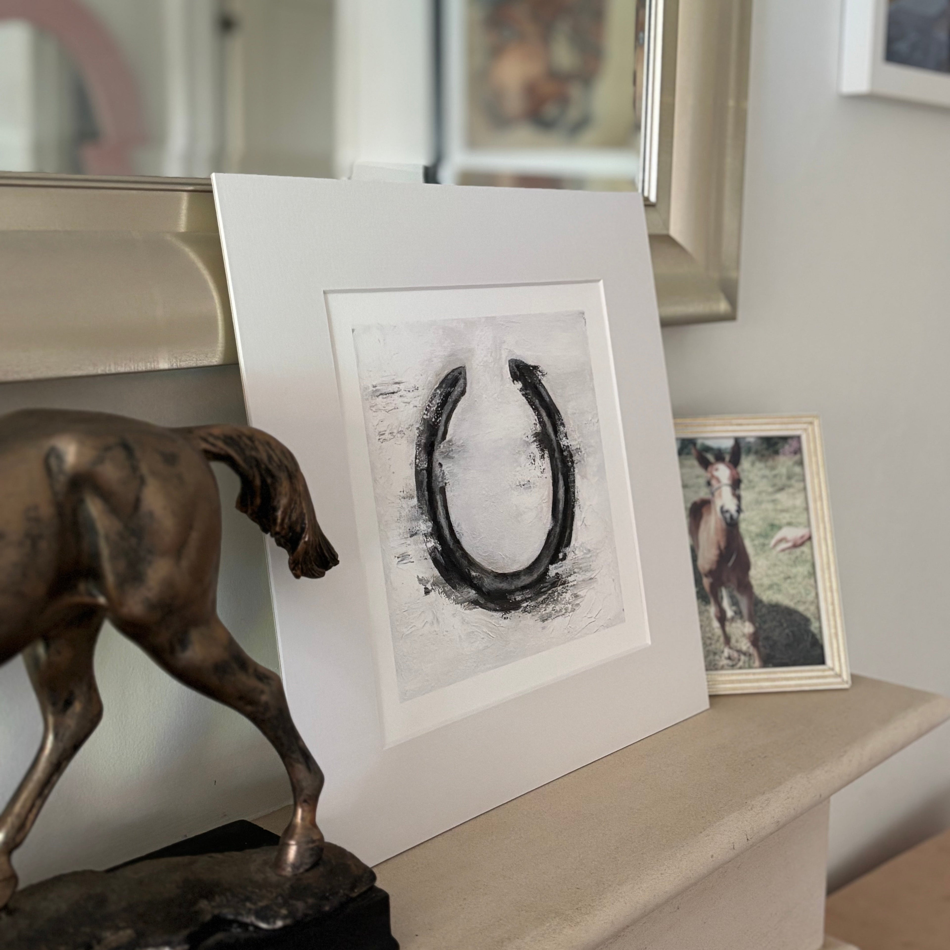 Framed artwork of a horseshoe on a mantelpiece with decorative items.