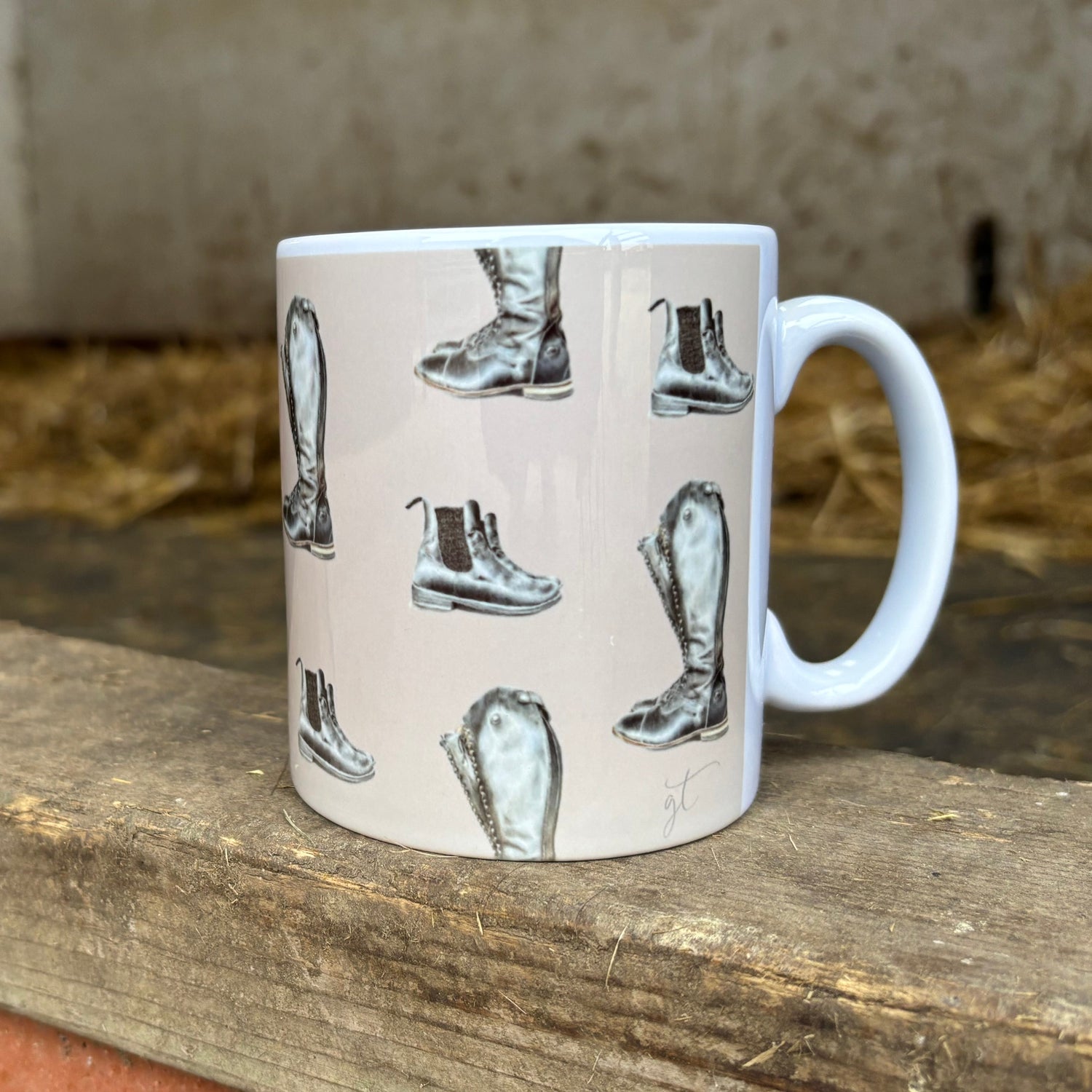White mug with black boot design on a wooden surface with hay in the background