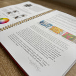 An open spiral-bound book lying on a wooden desk, showing printed text, small illustrations, and a red cover edge. A laptop is partially visible in the blurred background.
