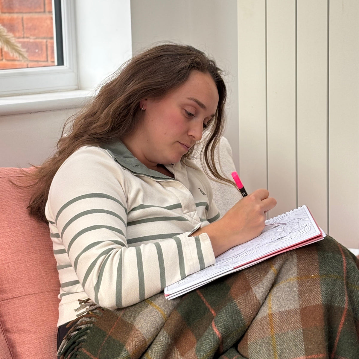 A young woman sits on a pink sofa, writing in a notebook with a pink pen while wrapped in a brown and green plaid blanket. She is wearing a cream and green striped long-sleeve top.