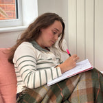 A young woman sits on a pink sofa, writing in a notebook with a pink pen while wrapped in a brown and green plaid blanket. She is wearing a cream and green striped long-sleeve top.