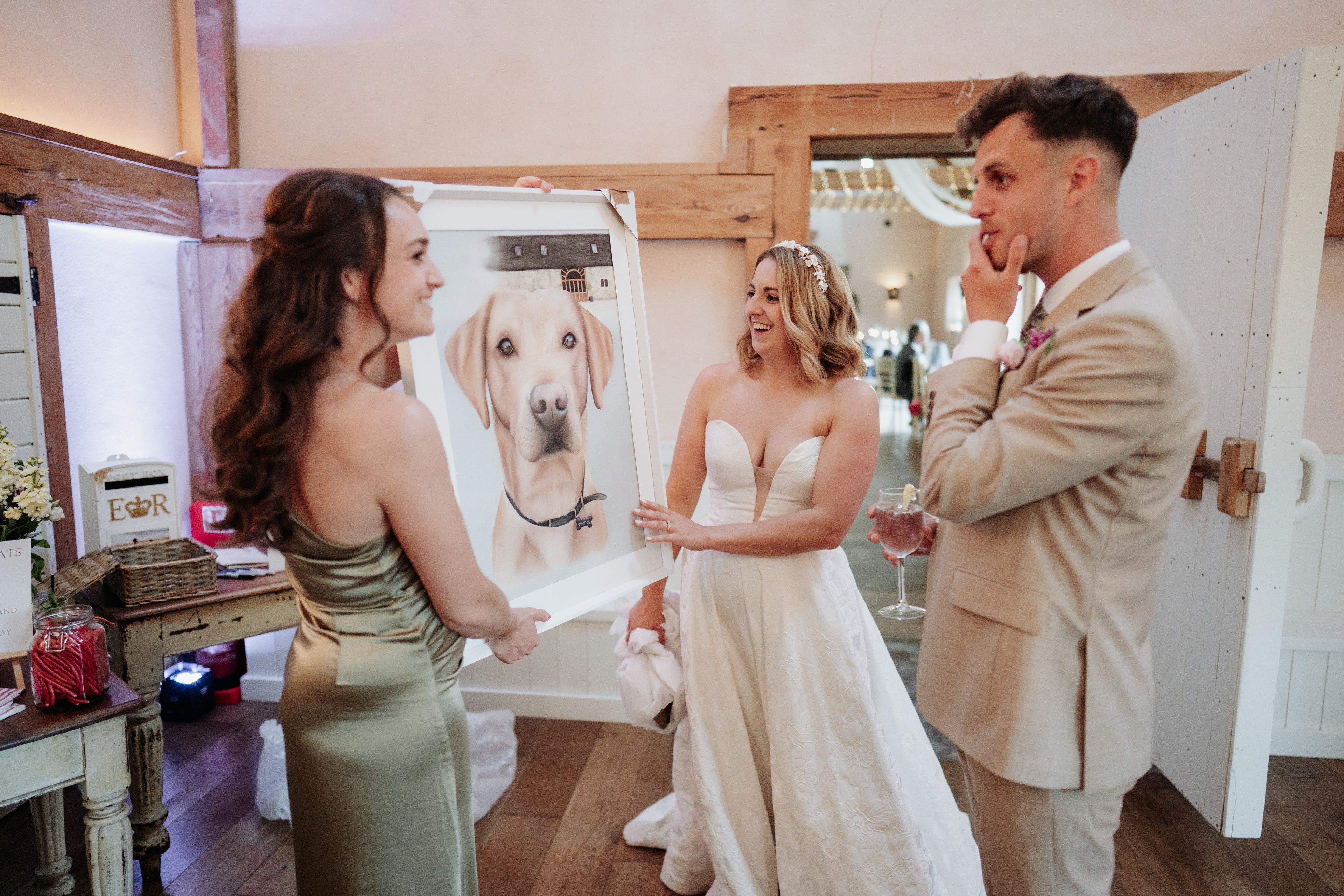 Woman in a wedding dress holding a portrait of a dog, with a man in a suit and another woman in a green dress in a room.